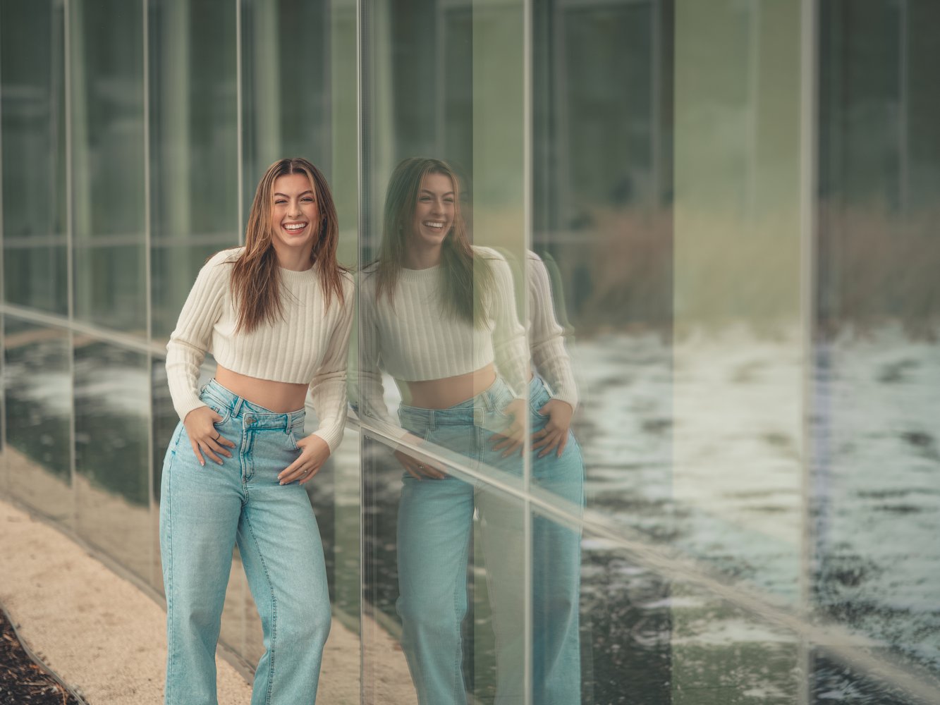 Big smile for Annie leaning against a wall of windows in her jeans and cropped ivory sweater for senior pictures